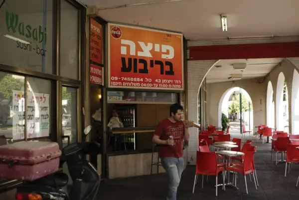 החזית של סניף פיצה בריבוע: שלט כתום, כיסאות אדומים ומרפסת מקושתת